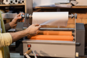 Close-up of a person's hands operating a industrial laminating machine. The machine is processing a roll of white laminating film over orange rollers. The background shows a workspace with shelves containing rolls of materials, creating a professional and organized environment for document or material lamination.