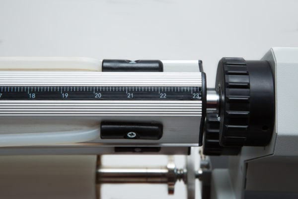 A close-up view of a laminator machine's adjustable tension control knob and measurement scale. The metallic roller and black dial are used to fine-tune the lamination process, ensuring precise application of laminating film. The scale is marked with measurements for accurate adjustments.